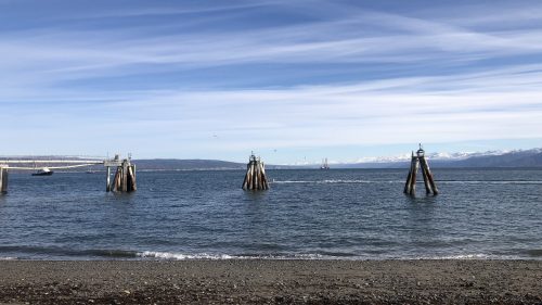 A serene coastal scene with wooden pilings in the water, mountains in the distance, and a clear blue sky.
