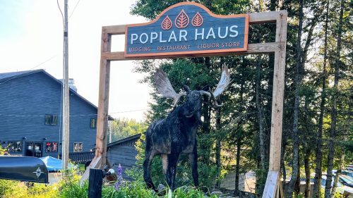Sign for Poplar Haus featuring a moose statue, surrounded by greenery and flowers, with buildings in the background.