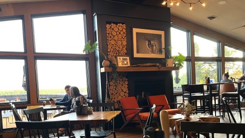 Cozy restaurant interior with large windows, wooden decor, and patrons enjoying their meals at tables.