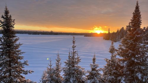 A serene winter landscape with snow-covered trees and a sunset over a frozen lake.