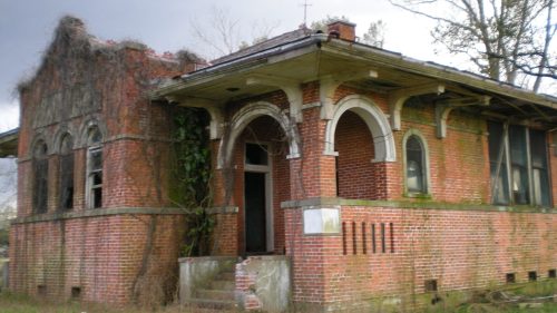 Abandoned brick house overgrown with vines, featuring arched windows and a deteriorating roof, set against a cloudy sky.