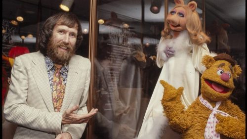 A man in a suit poses with Miss Piggy and Fozzie Bear puppets in a display setting.
