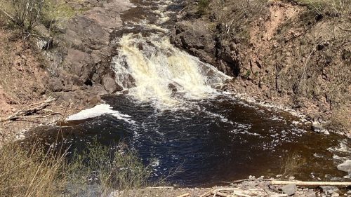 A serene waterfall cascades through rocky terrain, surrounded by lush green trees and a clear blue sky.