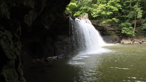 A serene waterfall cascades into a calm pool, surrounded by lush green trees and rocky cliffs.