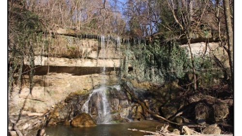 A serene waterfall cascading over rocky terrain, surrounded by lush greenery and trees under a clear blue sky.