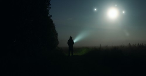The silhouette of a person at night holding a flashlights and looking at the sky with orb-like lights floating around. This is the Joplin Spooklight, a hidden legend in Missouri near Joplin.