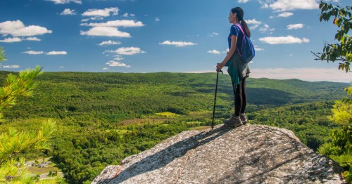A woman with a walking stick stands on a rock and looks out atop Monument Mountain in Great Barrington, MA.