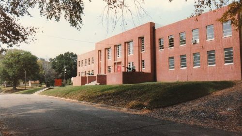 A brick school building with large windows, set on a grassy slope beside a quiet street. Trees frame the scene.
