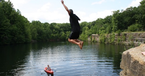 A man is in midair jumping into a lake surrounded by green trees while a red kayak waits in the water.