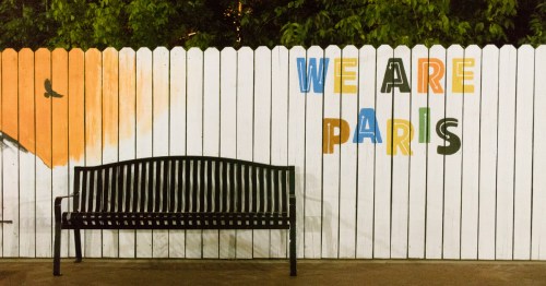 A black park bench in front of a white picket fence painted with the colorful words "WE ARE PARIS."