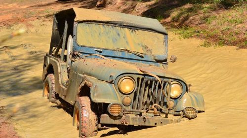 A muddy vintage jeep partially submerged in water, with a second vehicle visible in the background.