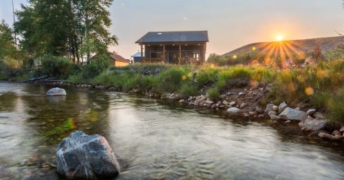 The sun begins to set behind a small rustic cabin along a slow river in Wyoming.
