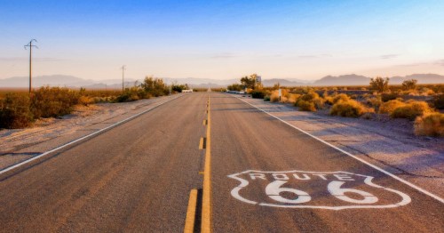 The Route 66 shield painted on a desert highway stretching toward distant mountains at sunrise.