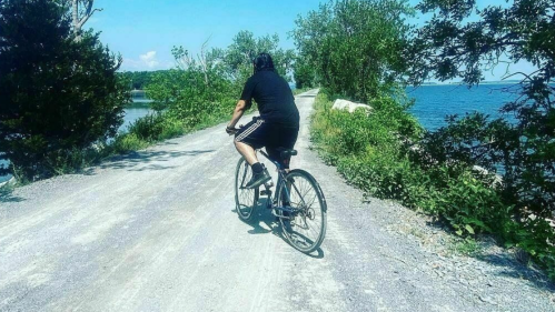 A person rides a bicycle along a gravel path by a lake, surrounded by trees and blue skies.