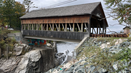 A wooden covered bridge spans a small dam, with rocks and greenery surrounding the area under a cloudy sky.