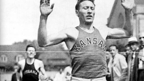 A runner in a Kansas tank top crosses the finish line, celebrating victory, with another competitor behind him.