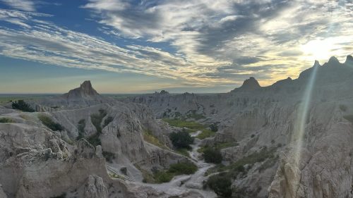 A scenic view of rugged, eroded rock formations under a cloudy sky at sunset, with green vegetation in the valley.