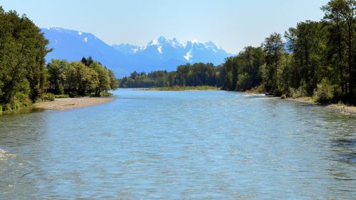 A serene river flows through a lush landscape, with snow-capped mountains in the background under a clear blue sky.