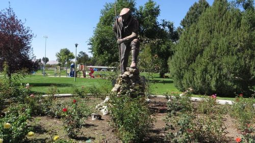 A statue of a worker in a garden, surrounded by rose bushes and trees, with a playground visible in the background.