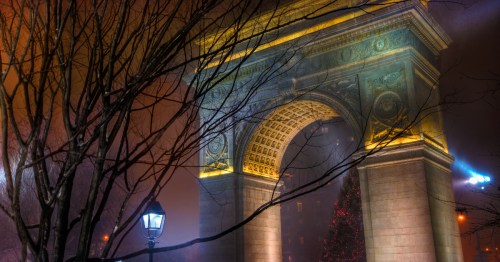 An eerie nighttime view of the Washington Square Arch in New York City's Washington Square Park. The arch is partially covered by bare tree branches.