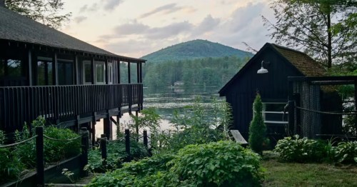 Black timber lakeside cottage on stilts beside Tully Pond, with a guest cabin and Tully Mountain behind.