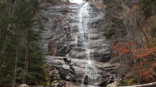 A cascading waterfall flows down a rocky cliff, surrounded by trees and autumn foliage.