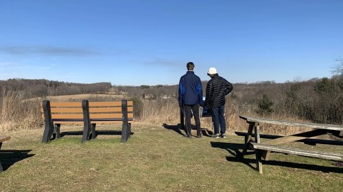 Two people stand on a grassy overlook, gazing at a scenic view with benches and picnic tables nearby.