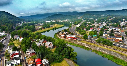Aerial view of Cumberland, Maryland, showing blue mountains in the distance and a river winding through with train tracks and homes.