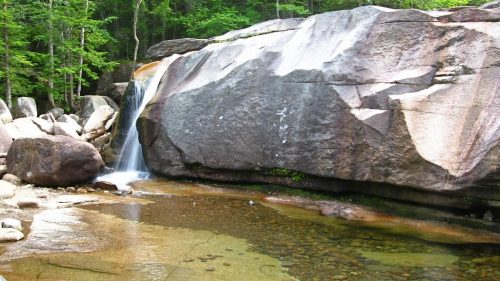 A serene waterfall cascades over a large rock into a clear pool, surrounded by lush green trees and boulders.