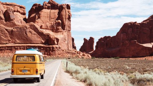 A yellow van drives along a road surrounded by towering red rock formations and desert landscape under a blue sky.