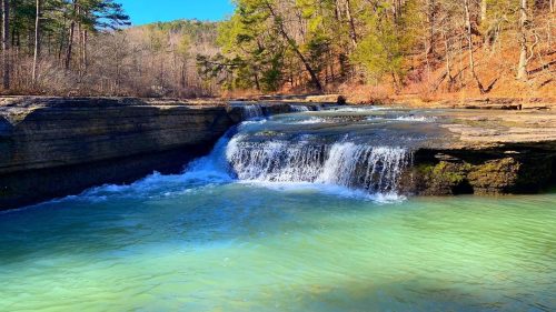 A serene waterfall cascades into a clear turquoise pool, surrounded by trees and rocky terrain under a bright blue sky.