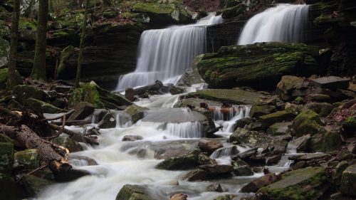 A serene waterfall cascades over moss-covered rocks, surrounded by lush greenery and flowing water.