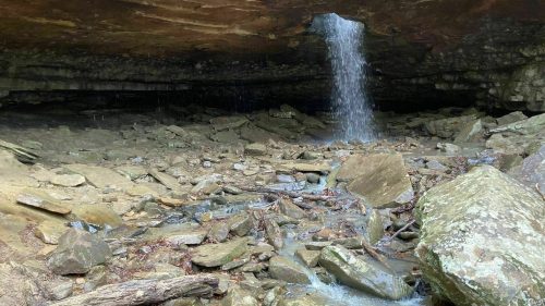 A small waterfall cascades from a rocky overhang into a stony creek bed surrounded by large boulders.