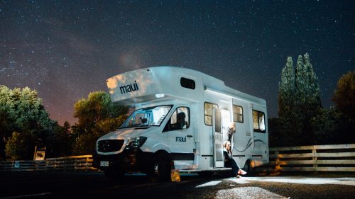 A camper van parked under a starry sky, with a person standing outside, surrounded by trees.