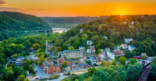 Sunset view of Harpers Ferry, WV, as seen from Maryland Heights. Historic town buildings with mountains and orange sky overhead.