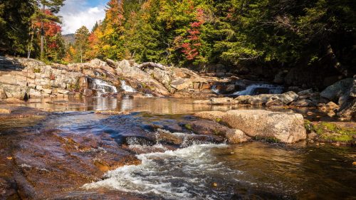 A serene river flows over rocks, surrounded by colorful autumn trees under a clear blue sky.