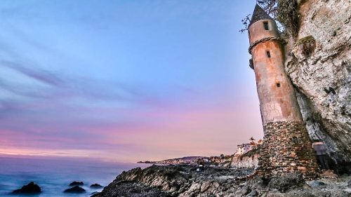 A coastal tower stands on rocky shore at sunset, with waves gently lapping against the stones and a colorful sky.