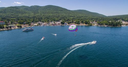 A boat with a parasailing balloon rides through the waters of Lake George, New York, on a sunny day with the town in the back.