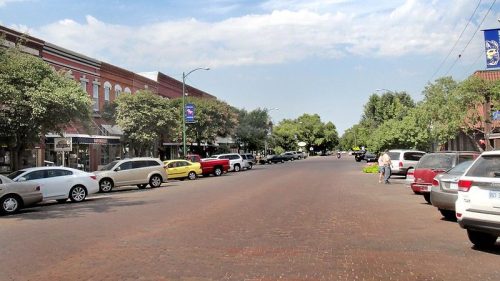 A wide street lined with trees and parked cars, featuring shops on either side under a blue sky.