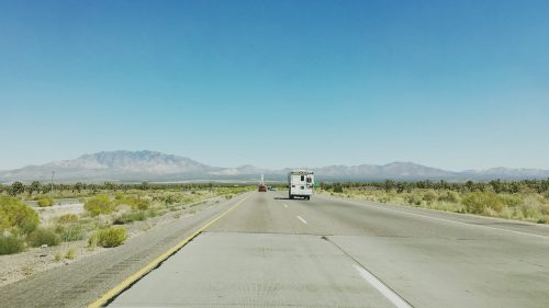 A clear highway stretches into the distance, flanked by desert vegetation and mountains under a blue sky.