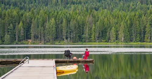 Two people sit in camping chairs at the end of a dock on a quiet northern Idaho lake.