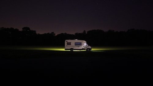 A camper van parked in a dark field, illuminated by its own lights under a starry night sky.