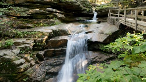 A serene waterfall cascades over rocky terrain, surrounded by lush greenery and a wooden walkway in a forested area.