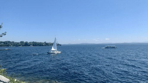A sailboat glides across a calm blue lake under a clear sky, with distant hills visible on the horizon.