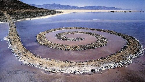 A spiral stone formation on a salt flat, surrounded by water and mountains in the background under a clear blue sky.