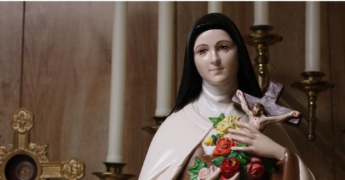 Statue of the Virgin Mary holding a rosary next to flowers and candle holders.