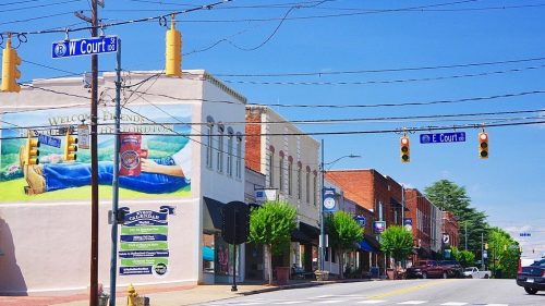 A sunny street scene featuring shops, traffic lights, and a mural in a small town. Trees line the sidewalk.