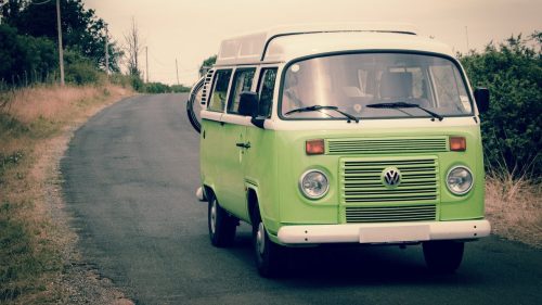 A vintage green Volkswagen van parked on a winding road surrounded by grass and trees.