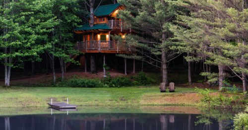 Illuminated Vermont treehouse rental glowing at twilight above a calm pond and dock framed by tall pines.