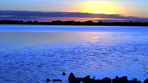 A serene lake at sunset, with vibrant blue and orange hues reflecting on the water and silhouettes of trees in the background.